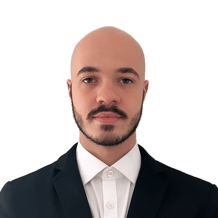 A bald man with a beard is wearing a black suit and white shirt, looking directly at the camera against a plain white background.