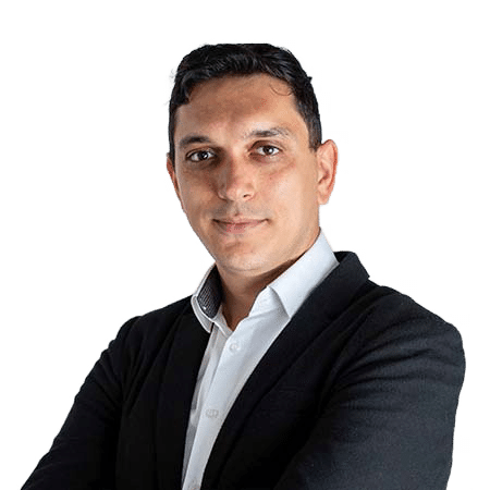 A person with short dark hair is wearing a white shirt and black jacket, looking directly at the camera with a neutral expression. Arms are crossed in front.